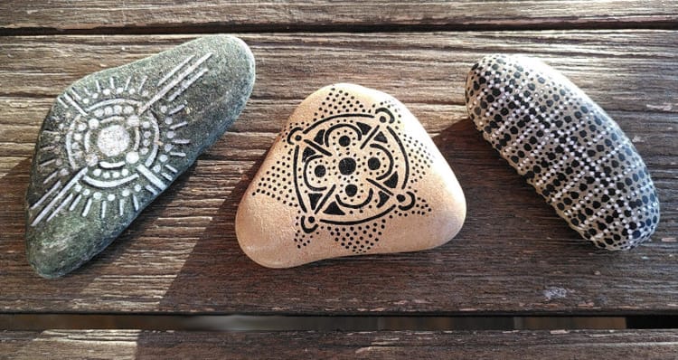 Trois cailloux décorés de points et de traits effectués aux feutres noirs ou blancs, disposés sur une table en bois