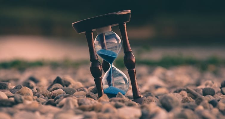 Tilted hourglass resting on small pebbles. Blue sand flows inside.